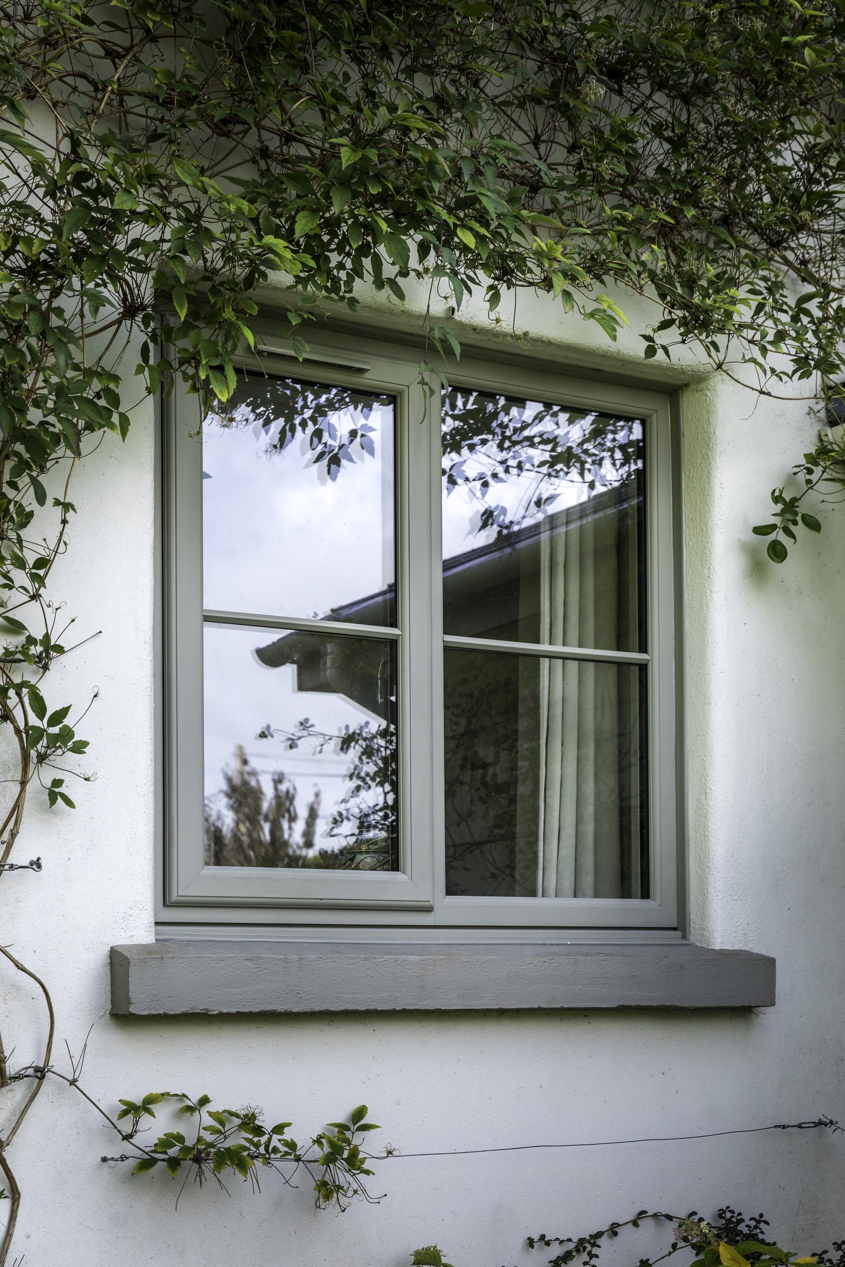 Sage green uPVC casement window with a horizontal astragal bar, fitted into a white cottage exterior wall surrounded by climbing green ivy.