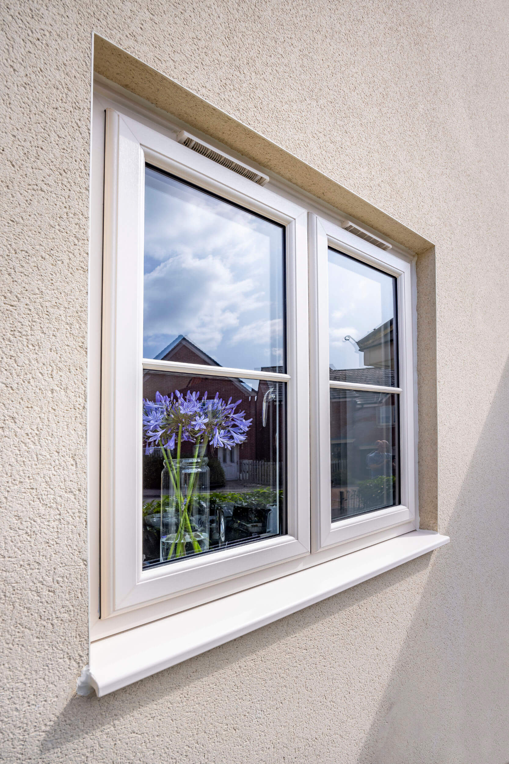 Angled close-up of a Cream uPVC casement window with internal astragal bars and trickle vents built into a beige textured rendered exterior wall